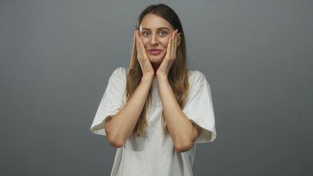 Young woman in white t shirt pressing hands to her cheeks with open mouth in gray studio looking at camera with long hair; surprise.