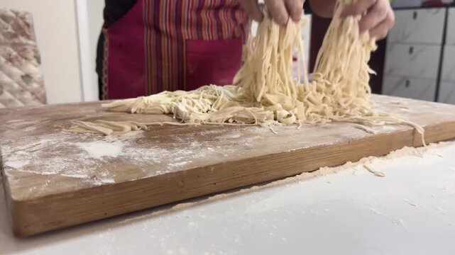 Preparing fresh noodles from scratch at home by shaping and cutting dough on a floured wooden board, showcasing traditional homemade cooking in a kitchen setting.