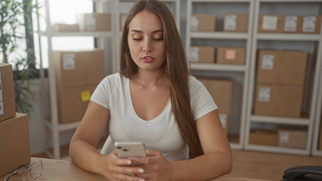Woman holding smartphone at shipping office desk with stacked parcel boxes, visible labels and eyeglasses on table; small business planning calm.