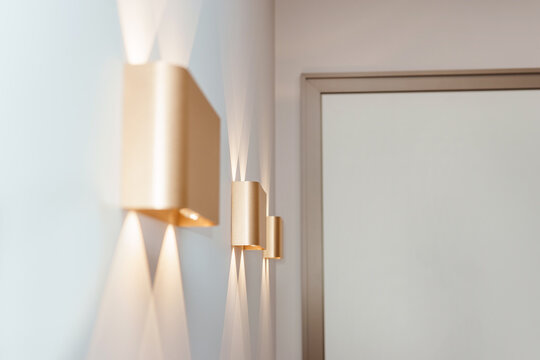 Modern wall lamps with glowing light beams in minimalist home interior. Stylish golden sconce on white wall for luxury house decor. Contemporary lighting design and architecture element.