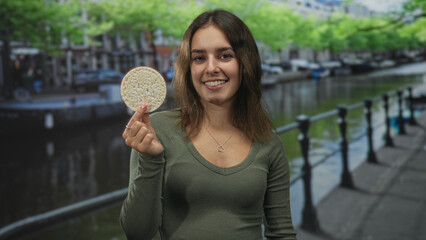 Young hispanic woman smiling and holding rice cake by canal railing on street in amsterdam, near...