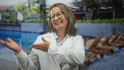 Fototapeta premium Hispanic woman in white coat gesturing with hands and smiling in studio by poolside lounge chairs; confidence reassurance.