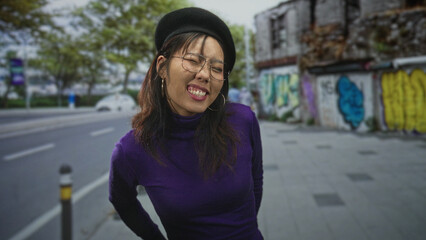 Fototapeta premium Woman wearing beret and glasses tilts head with eyes closed and hands behind back on city street beside graffiti wall, purple turtleneck visible; playful ease.