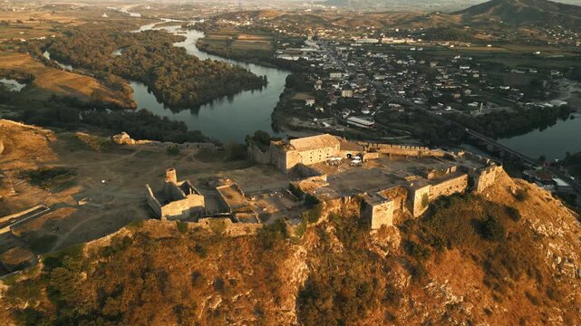 Aerial drone orbiting left around Rozafa Castle at sunset, golden light over fortress ruins with river valley and Shkoder city in warm evening atmosphere