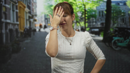Fototapeta premium Woman facepalming with hand over forehead, showing watch and rings, standing in a narrow city street with bikes and buildings in view; frustration.