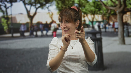 Fototapeta premium Woman in white blouse points finger to camera on street with trees and trashcan nearby; playful confidence.
