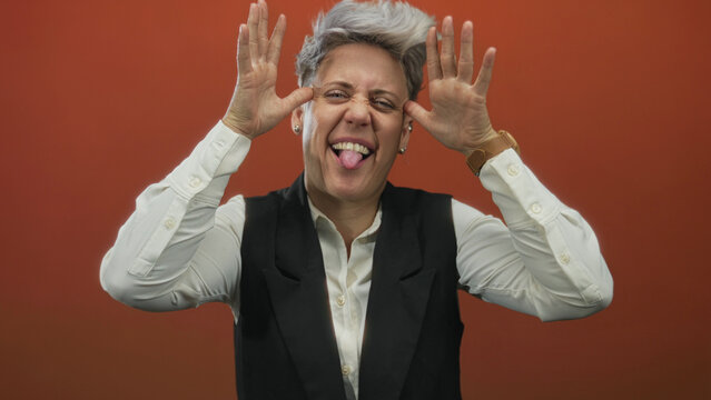 Woman framing her face with both hands and sticking out her tongue in a studio with an orange wall; playfulness.