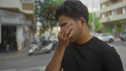 Fototapeta premium Man with hand covering face and visible fingers over eyes on street near parked scooters and multistory building; stress reflection.