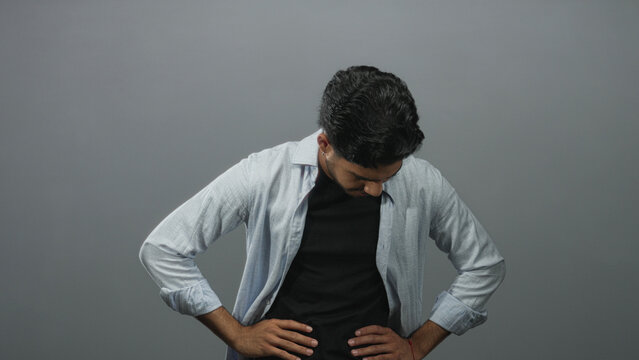 Man touching his abdomen and adjusting an open shirt while looking down in a gray studio setting; discomfort.