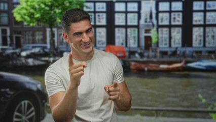Fototapeta premium Man points finger with both hands on a canal street in amsterdam near boats and cars, smiling in a white t shirt; playful confidence.