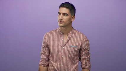 Obraz premium Man points finger to left in studio with purple backdrop, wearing striped shirt and leaning forward; surprise attention.
