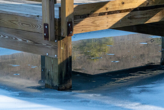 Sunlit Wooden Dock Structure with Strong Shadows Reflected in Partially Frozen Water and Ripples, Featuring Wood Grain and Metal Fasteners in Winter