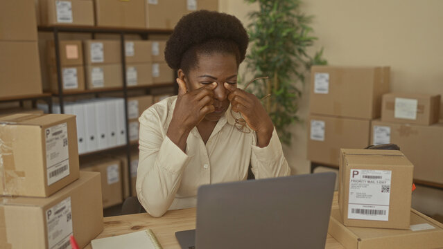 Woman removes glasses rubbing eyes at warehouse desk with shipping boxes and laptop; fatigue resilience entrepreneurship.