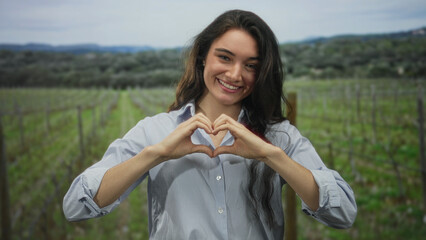 Naklejka premium Young hispanic woman smiling in a green countryside field forms heart shape with fingers toward camera; love.