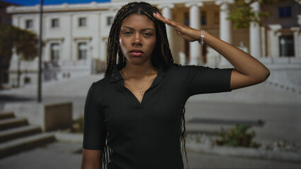 Fototapeta premium African american woman salutes with hand to forehead in front of building, wearing black top and cross necklace, braids visible; respect duty determination.