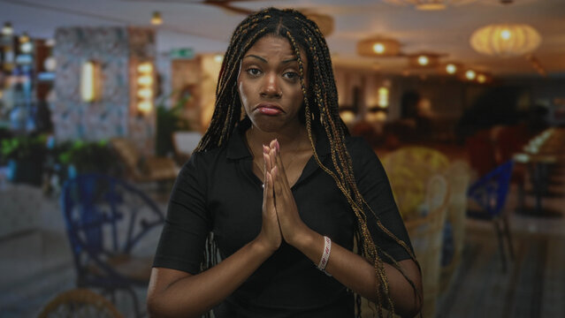 Woman pressing hands together with palms visible and braided hair in a hotel building; pleading apology request.