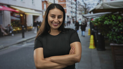 Fototapeta premium Young hispanic woman with crossed arms showing forearms and smiling on a busy city street cafe setting; confidence empowerment.