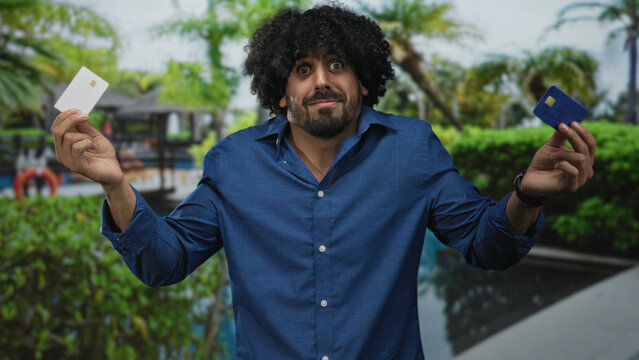 Man holding two creditcards in both hands at a building pool area showing a hesitant shrug; financial uncertainty.