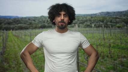 Naklejka premium Man with curly hair and beard, hands on hips pose, wearing white tshirt with chest visible in forest vineyard rows; surprise.