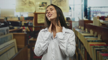 Young hispanic woman with hand on neck in library showing throat pain and strained expression amid bookshelves and records  discomfort. © Krakenimages.com