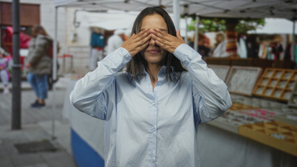 Fototapeta premium Woman clasping fists and covering eyes in a playful grin at street market stalls; joy playful surprise.