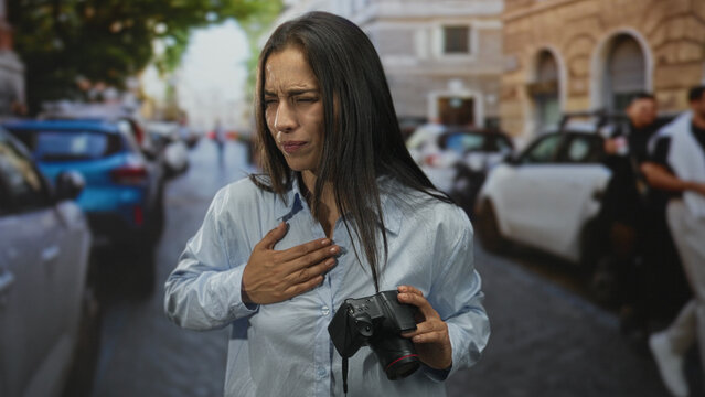 Young hispanic woman clutching chest holding dslr camera on busy city street after sudden chest pain for heartattack; panic concern.