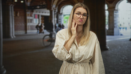 Obraz premium Blonde woman with glasses hand on cheek holding jaw for toothache under a stone building arcade while a cyclist rides past; discomfort.