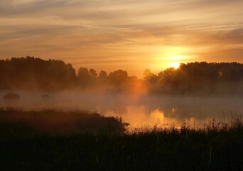 Obraz premium The sun rising over a misty forest and lake, creating a warm orange glow. Stunning morning landscape showcasing the beauty of Masurian nature.
