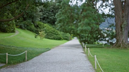 Fototapeta premium Blurred park path with defocused trees, lawn, rope posts and a gravel walkway leading into soft distance; background backplate copyspace calm.