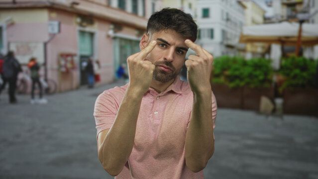 Young hispanic man in pink polo shirt raises middle fingers while walking on urban street; defiance.