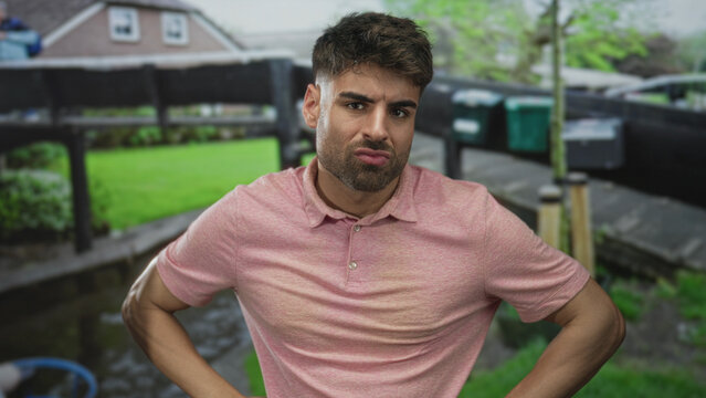 Man in pink polo shirt standing with hands on hips and raised eyebrow in front of building; defiance.