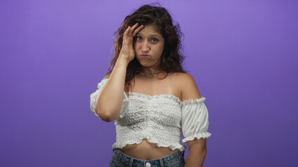 Young hispanic woman wearing white crop top with hand to forehead and exposed midriff in purple studio, uneasy expression and squint; introspection fatigue.