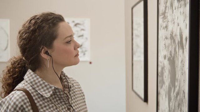 Side view slowmo shot of focused young Caucasian female gallery visitor wearing audio guide earphones looking at framed abstract paintings on exhibition wall