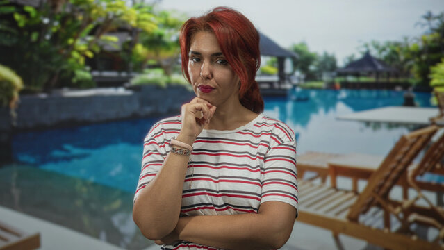 Woman with hand on chin and folded arms at poolside building with lounge chairs and tropical plants; thoughtful reflection.
