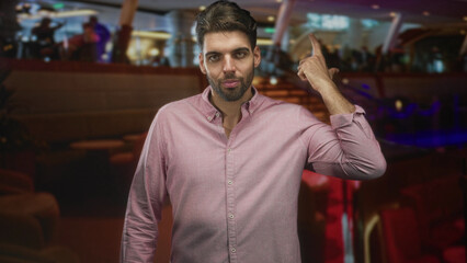 Fototapeta premium Man with beard beckoning with hand in airport lounge, wearing pink shirt and standing near seats and railing; assertion confidence.