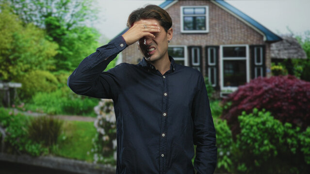 Man presses forehead with hand while squinting in front of a suburban building garden, wearing a blue shirt and earring; headache distress.