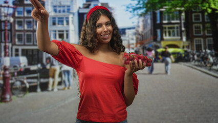 Obraz premium Hispanic teenage woman holding strawberries in hand on street market, red off shoulder blouse and headband visible; joy summer freshness.