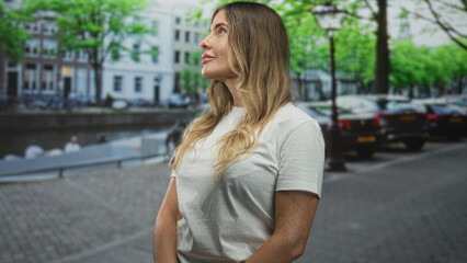 Fototapeta premium Middleaged hispanic woman face smiling and looking up on street by a canal with parked cars, lamp post, trees and riverside buildings; contentment.