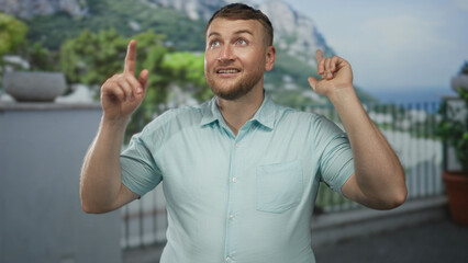 Fototapeta premium Man in light blue shirt with beard and bare forearms points both index fingers up on building balcony, smiling and looking upward at sky; hope future.