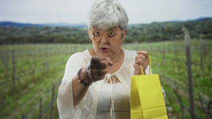 Naklejka premium Senior woman with gray hair and glasses points finger while holding a yellow shopping bag in a vineyard among vine rows; assertive invitation.