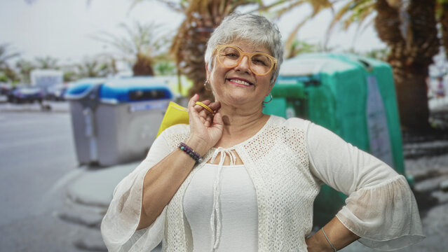Woman senior with gray hair wearing glasses and crochet cardigan, holding yellow shopping bag with hand, smiling on street; joy confidence ease.