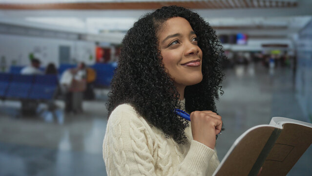 Woman smiling and holding pen while jotting notes in notebook at busy airport terminal with blurred travelers; planning future.