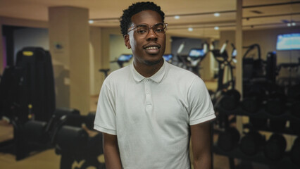 Man wearing glasses and a white polo shirt stands smiling by a weight rack in a modern gymnasium  confidence. © Krakenimages.com