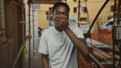 Fototapeta premium Man wearing white polo shirt covers mouth near scaffolding and orange netting on street construction site; surprise.