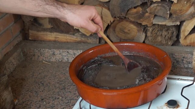 Hand stirring a thick, dark lamprey fish stew with a wooden spoon in a traditional clay pot over a gas stove. Rustic kitchen background with firewood.