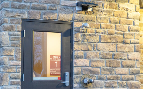 Modern building exterior featuring a textured natural stone brick facade, a dark grey fire exit door with safety signage, contemporary LED outdoor lighting, and a prominent fire department connection.