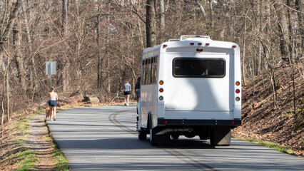 White shuttle bus drives on a curving asphalt road through a bare winter forest as two women walk, one with her dog, on parallel paths, illustrating outdoor activity and serene travel. © Michael