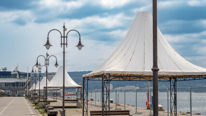Coastal promenade featuring white peaked tents and vintage-style street lamps. A calm seaside walkway under a cloudy sky, perfect for travel and urban leisure themes © John_Doo78