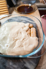 Fototapeta premium Homemade Tiramisu preparation in a glass baking dish, layering creamy mascarpone over espresso-soaked ladyfingers, a traditional Italian dessert in progress.