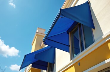 Fototapeta premium Blue fabric awnings extend from a building facade offering shade and style. Bright sunny day with clear blue sky and scattered clouds. The awnings provide cool comfort.